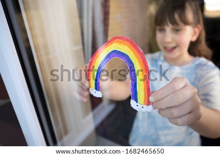 Girl Putting Picture Of Rainbow In Window At Home During Coronavirus Pandemic To Entertain Children