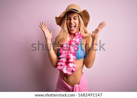 Young beautiful blonde woman on vacation wearing bikini and hat with hawaiian lei flowers celebrating mad and crazy for success with arms raised and closed eyes screaming excited. Winner concept