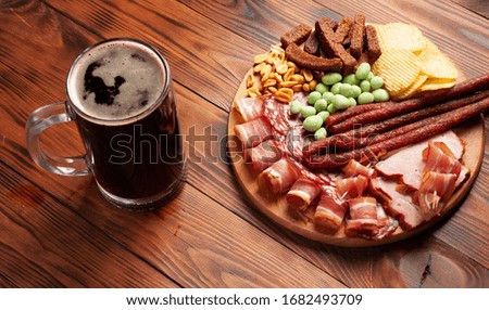 Mug of beer and assortment of snacks on a wooden table. Sausage, salami, ham, bacon. Beer salty snack, chips, wasabi nuts, salted peanuts, croutons.