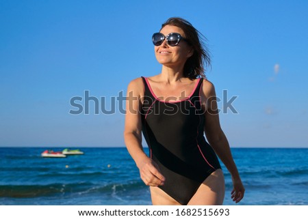 Mature smiling woman in swimsuit with sunglasses walking along beach. Beauty, health, body, relaxation for middle-aged people. Blue sky, sea with waves background, copy space