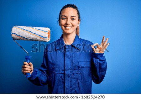 Young beautiful blonde painter woman with blue eyes painting wearing uniform using roller doing ok sign with fingers, excellent symbol