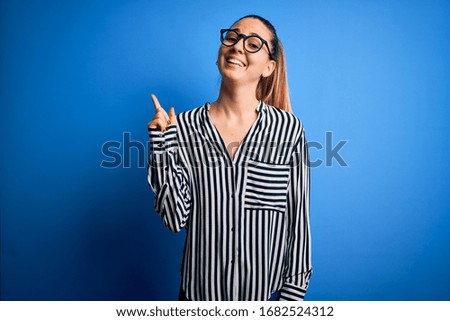 Beautiful blonde woman with blue eyes wearing striped shirt and glasses over blue background with a big smile on face, pointing with hand and finger to the side looking at the camera.