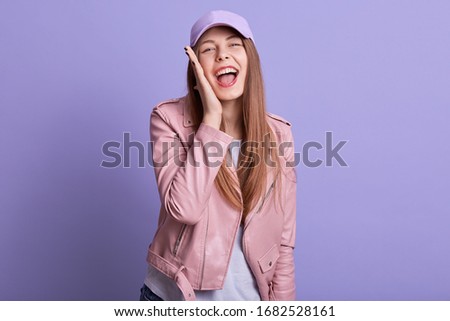 Picture of attarctive woman dresses leather pink jacket and cap cavering ear with palm screaming something, expressing happyness, looks directly at camera, being in good mood. People emotions concept.
