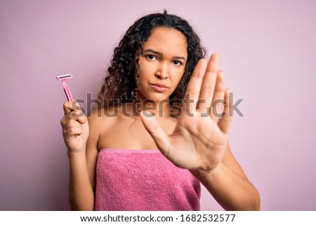 Young beautiful woman with curly hair wearing shower towel holding depilation razor with open hand doing stop sign with serious and confident expression, defense gesture