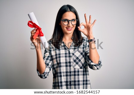Young brunette student woman with blue eyes holding university diploma over isolated background doing ok sign with fingers, excellent symbol