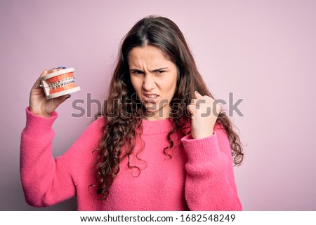 Beautiful woman with curly hair holding plastic teeth with dental braces over pink background annoyed and frustrated shouting with anger, crazy and yelling with raised hand, anger concept