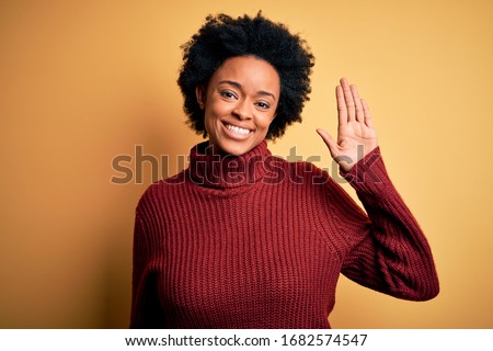 Young beautiful African American afro woman with curly hair wearing casual turtleneck sweater Waiving saying hello happy and smiling, friendly welcome gesture