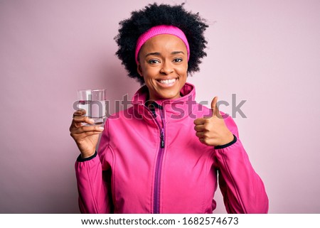 Young African American afro sportswoman with curly hair drinking glass of water happy with big smile doing ok sign, thumb up with fingers, excellent sign