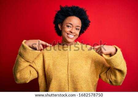 Young beautiful African American afro woman with curly hair wearing casual yellow sweater looking confident with smile on face, pointing oneself with fingers proud and happy.
