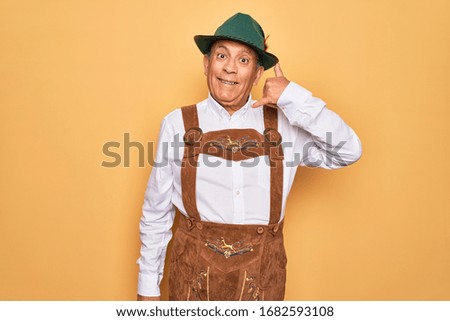 Senior grey-haired man wearing german traditional octoberfest suit over yellow background smiling doing phone gesture with hand and fingers like talking on the telephone. Communicating concepts.