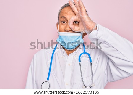 Senior hoary doctor man wearing medical mask and stethoscope over pink background with happy face smiling doing ok sign with hand on eye looking through fingers