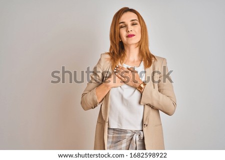 Redhead caucasian business woman standing over isolated background smiling with hands on chest with closed eyes and grateful gesture on face. Health concept.