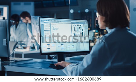 Over the Shoulder Shot: Female IT Scientist Uses Computer Showing System Monitoring and Controlling Program. In the Background Technology Development Laboratory with Scientists, Engineers Working Royalty-Free Stock Photo #1682600689