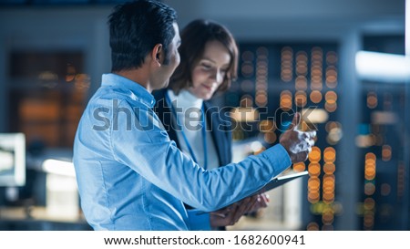 In Technology Research Facility: Female Project Manager Talks With Chief Engineer, they Consult Tablet Computer. Team of Industrial Engineers, Developers Work on Engine Design Using Computers Royalty-Free Stock Photo #1682600941