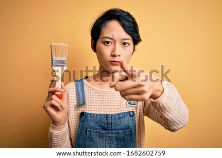 Young beautiful chinese woman painting holding paint brush over isolated yellow background pointing with finger to the camera and to you, hand sign, positive and confident gesture from the front