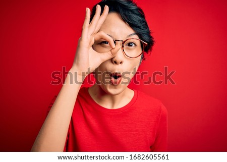 Young beautiful asian girl wearing casual t-shirt and glasses over isolated red background doing ok gesture shocked with surprised face, eye looking through fingers. Unbelieving expression.