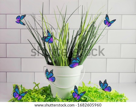 White metal bucket full of artificial grass sitting on a bed of green paper shreds with a a white subway tile background, swarmed with blue and purple butterflies.  Spring home decor.