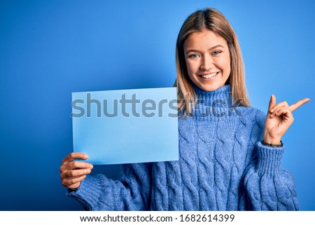 Young beautiful woman holding banner standing over isolated blue background very happy pointing with hand and finger to the side