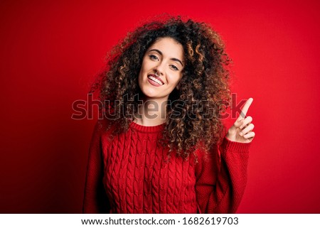 Young beautiful woman with curly hair and piercing wearing casual red sweater with a big smile on face, pointing with hand finger to the side looking at the camera.