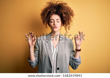 Young beautiful businesswoman with curly hair and piercing wearing elegant jacket relax and smiling with eyes closed doing meditation gesture with fingers. Yoga concept.