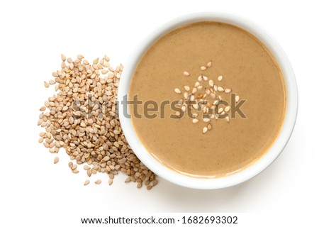 Tahini in a white ceramic bowl sprinkled with sesame seeds next to a pile of sesame seeds isolated on white. Top view. Royalty-Free Stock Photo #1682693302