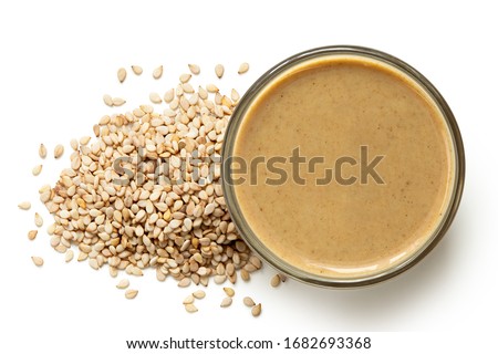 Tahini in a glass bowl next to a pile of sesame seeds isolated on white. Top view. Royalty-Free Stock Photo #1682693368