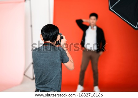 back view of male asian professional photographer taking picture of male model in red isolated background with studio lighting umbrella soft box, representing photographer studio lighting concept