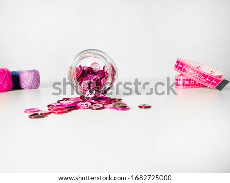 Glass jar full of clothing pink buttons, measuring tape and threads on white background. Sewing and craft concept.