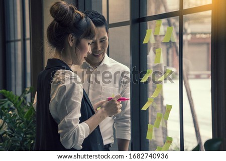 Asian creative business female and male thinking with note and sticking at contact sheet on glass wall in office.