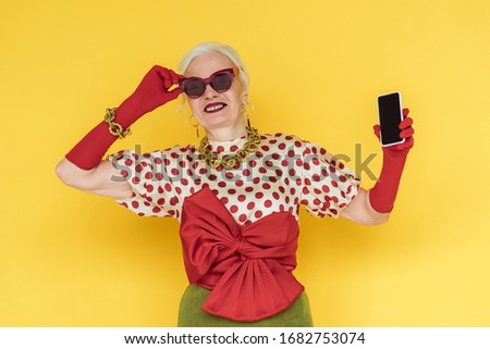 Stylish senior woman smiling and holding smartphone with blank screen on yellow background