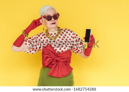 Fashionable senior woman pointing with finger on head and holding smartphone isolated on yellow