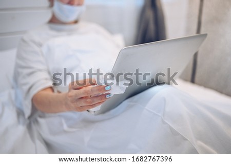 Unrecognizable woman in face mask in bedroom during coronavirus isolation home quarantine cleaning laptop by hand sanitizer, using cotton wool with alcohol to wipe to avoid contaminating with COVID-19
