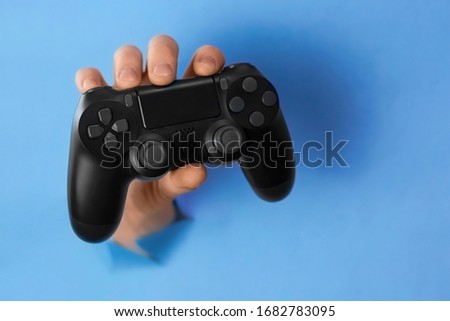 Men's hand holds video game controller through a hole in torn blue paper wall. Horizontal picture with copy space and selective focus. Stay home concept 