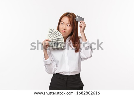 Portrait of hesitant and unsure young asian woman having doubts, holding money and credit card, scratch head uncertain look away thoughtful, pondering decision, need buy something expensive