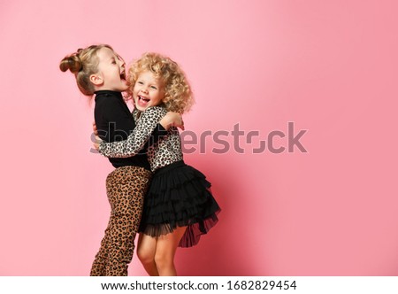 Two happy kids blonde girls best friends sisters in leopard print clothes pants and sweater are hugging laughing over pink background with free copy space