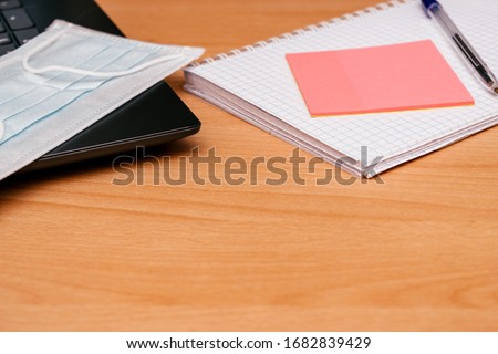 Concept work from home.Protective medical mask on the laptop,pen, notebook and pink stickers on the wooden table.Selective focus.Copy space for text,top view,close up