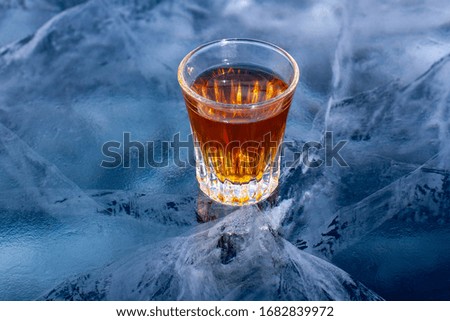 On the clear ice of the lake is a glass of whiskey. Chilled whiskey. Blue ice with beautiful deep white cracks. Top view from the side. Advertising alcohol products. Horizontal.