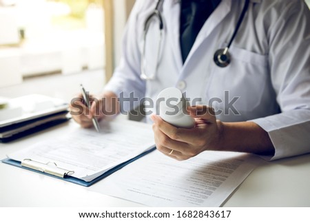Confident doctor man holding a pill bottle and writing while talking with senior patient and reviewing his medication at office room. Royalty-Free Stock Photo #1682843617
