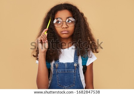 Clever African American kid in glasses scratching head with pencil and looking away while thinking over homework assignment against beige background Royalty-Free Stock Photo #1682847538