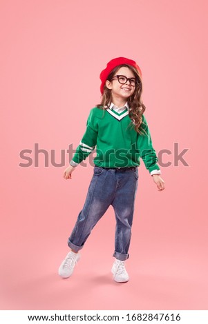 Full length cute little girl in stylish clothes and red beret smiling and looking away against pink background