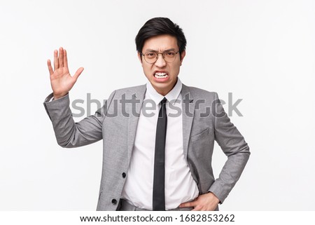 Waist-up potrait of angry, outraged asian businessman scolding employees, shouting and raising hand aggressive, grimacing with hate, being furious standing in suit over grey background