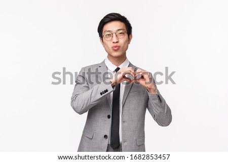 Waist-up portrait of handsome romantic male office worker in grey suit, showing heart sign and fold lips in kiss, looking camera, express like or sympathy, standing white background