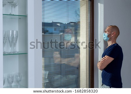 Home quarantine (self-isolation) because of the Coronavirus disease (COVID-19). Infected man with the ear-loop mask on his face looks out of the window. Man with protective face mask. Infection theme. Royalty-Free Stock Photo #1682858320