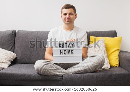 Young man sitting on the sofa in his living room holding lightbox with message Stay Home. Help spreading coronavirus Covid-19, quarantine, safety