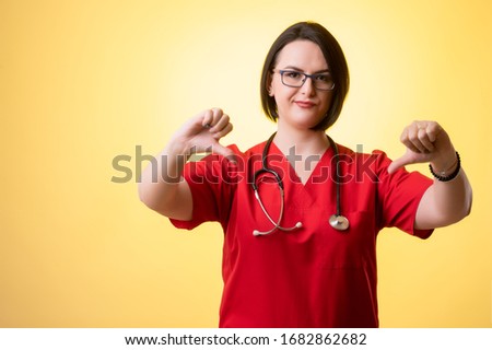 Portrait of beautiful woman doctor with stethoscope wearing red sctubs, showing double dislike posing on a yellow isolated background.