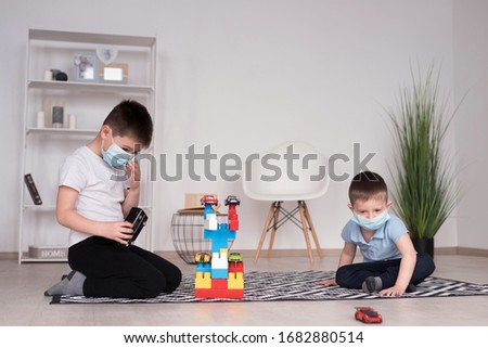 Two boys in medical masks play as a children's designer and toy cars  on the floor of the children's room. Covid-19. Healthy childhood concept. Coronovirus protection. Home insulation. Happy brothers
