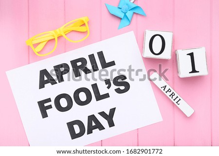 Text April Fool's Day with wooden calendar, eyeglasses and paper windmill on pink background