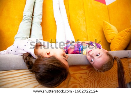 Beautiful two preteen girls sit in home  on color yellow sofa. Tween sisters chatting in a room at the company, lies upside down.