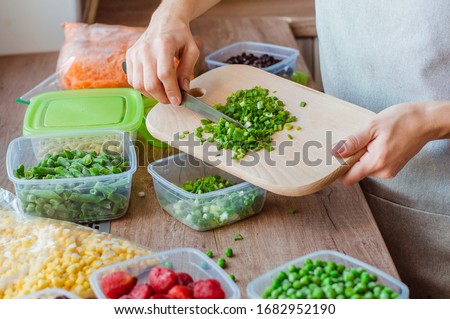 Close up of woman cutting green onion for freezing in the plastic food box on the wooden table. Plastic food boxes with another fresh vegetables and berries around it.
