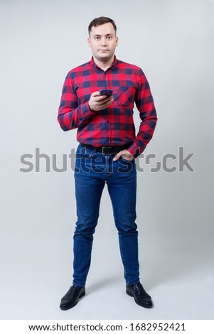 Young sincere man of 30 years old poses in a red checked shirt and jeans with a smartphone in his hand. Growth portrait on a gray background.